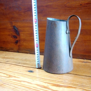 May include: A rusty metal pitcher with a handle. The pitcher is a light gray color and has a slightly conical shape.