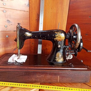 May include: A vintage black sewing machine with gold accents and a wooden base. The machine has a hand crank and a needle.