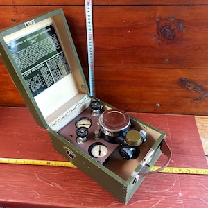 Puede incluir: Una caja de madera verde con una tapa con bisagras y un asa de cuero. La caja contiene un conjunto de cuatro indicadores y un cilindro de metal con una tapa negra. La tapa tiene una hoja de instrucciones impresa con el texto "Инструкция по пользованию прибором КИС".