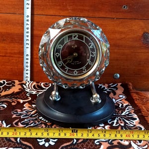 May include: A round table clock with a faceted clear glass frame, a dark brown dial, and silver-toned numerals. The clock is mounted on a black oval base with two silver metal supports. A measuring tape shows the clock is approximately 6 inches wide.
