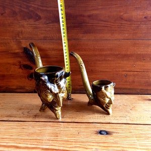 May include: Two ceramic pipes shaped like devil faces. The pipes are brown and green with black details. The pipes are on a wooden surface.