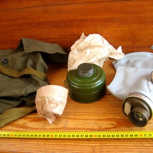 Op de afbeelding: Een groene gasmaskerfilter, een grijs gasmasker, een groene canvas tas met een band en een bruine papieren zak op een houten oppervlak. Een gele meetlint ligt op de voorgrond.