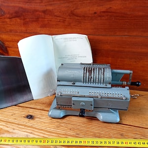 May include: A gray mechanical calculator with a lever on the right side. The calculator has a paper roll holder and a series of numbered dials. The calculator is labeled "AFRENOMETP" and "Феликс".