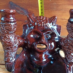 May include: A brown ceramic candle holder shaped like a devil's head with two candle holders on either side. The candle holder has a glossy finish and intricate details.