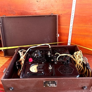 Puede incluir: Un dispositivo de prueba eléctrica negro vintage con un dial, perillas y un botón rojo, alojado en una caja de madera marrón. La caja está abierta, revelando los componentes y el cableado del dispositivo. Se ve una cinta métrica amarilla.