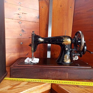 May include: A vintage black sewing machine with gold accents and a wooden base. The machine is sitting on a wooden surface with a measuring tape.