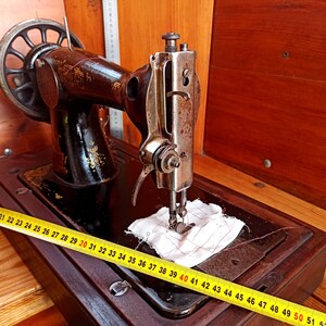 May include: A vintage sewing machine with a wooden base and a metal body. The machine has a hand crank and a needle that is threaded with white thread. The machine is sitting on a wooden table with a yellow measuring tape.