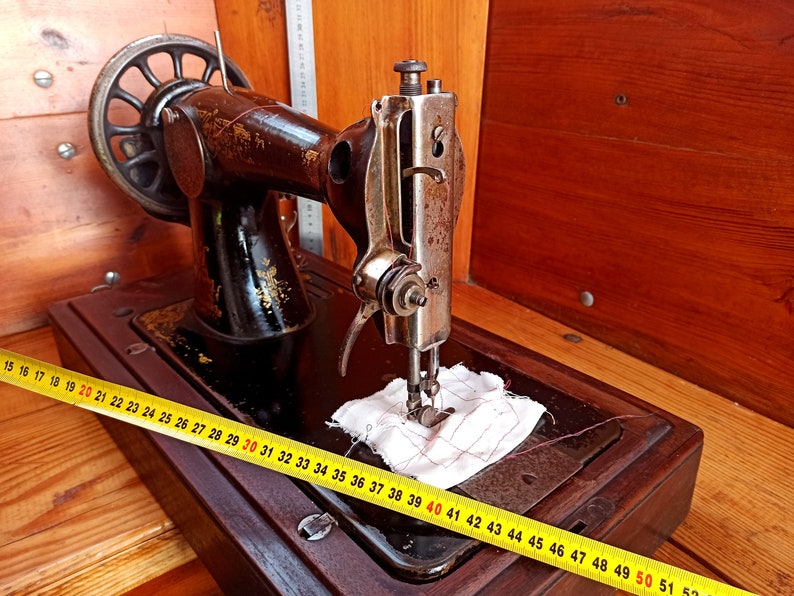 May include: A vintage sewing machine with a wooden base and a metal body. The machine has a hand crank and a needle that is threaded with white thread. The machine is sitting on a wooden table with a yellow measuring tape.