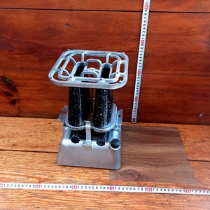 May include: A vintage silver-coloured camping stove with a black metal grate and two black fuel canisters. The stove is sitting on a wooden surface.