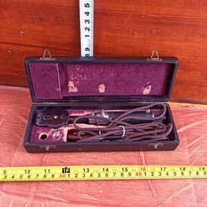 May include: An open, vintage black leather case with a maroon velvet interior. Inside, a black and silver instrument with a braided cord and a circular piece is visible. A ruler shows measurements in inches.