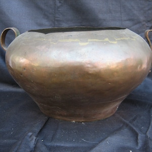 May include: A large, copper pot with two handles. The pot has a rounded bottom and a wide, open top. The copper has a warm, aged patina.