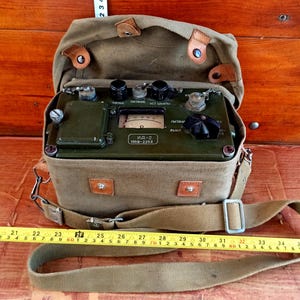 May include: An olive green vintage radiation detector, model ИД-2, inside a canvas bag with a shoulder strap. The detector has dials, knobs, and a meter. The bag is open, revealing the device's controls and a label with the text "ИД-2 1959-2268".