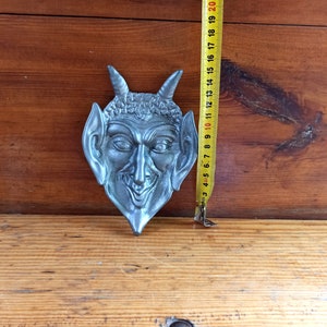 Puede incluir: Una escultura de pared de metal plateado con forma de cara de diablo con cuernos y una sonrisa traviesa.