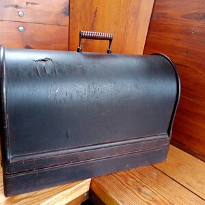 May include: A vintage wooden sewing machine case with a dark brown finish and a wooden handle. The case has a worn and weathered appearance.