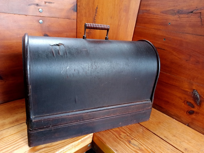 May include: A vintage wooden sewing machine case with a dark brown finish and a wooden handle. The case has a worn and weathered appearance.