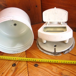 May include: White electric meter with a round base and a hinged lid. The lid is open and the meter face is visible. The meter face has a needle and a scale with markings for + P C. The meter is on a wooden surface.