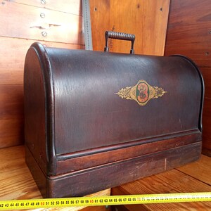May include: A vintage wooden sewing machine case with a dark brown finish. The case has a hinged lid with a metal handle and a decorative gold and black label with the number "3" on it.