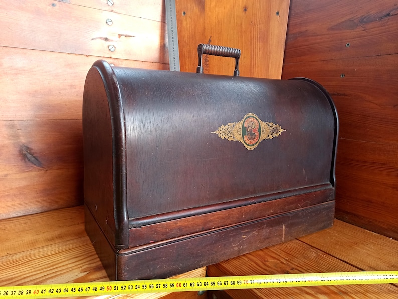 May include: A vintage wooden sewing machine case with a dark brown finish. The case has a hinged lid with a metal handle and a decorative gold and black label with the number "3" on it.