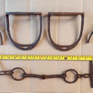 May include: A collection of antique iron equestrian equipment including two stirrups, two spurs, and a bit, all featuring a heavily rusted, textured brown finish. A yellow measuring tape is positioned horizontally, showing the items span 26 inches.
