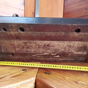 May include: The underside of a wooden box with three holes drilled into the surface. The box is dark brown and has a worn, aged appearance. A yellow measuring tape is visible in the bottom right corner of the image.