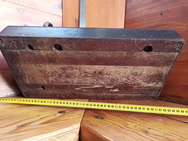 May include: The underside of a wooden box with three holes drilled into the surface. The box is dark brown and has a worn, aged appearance. A yellow measuring tape is visible in the bottom right corner of the image.