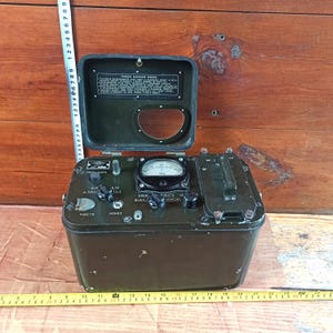 Puede incluir: Un dispositivo de detección de radiación de metal verde oliva con tapa abatible, medidor analógico y texto en cirílico. Una cinta métrica vertical muestra 24 cm de altura y una horizontal 24 cm de ancho. Incluye perillas e interruptores.