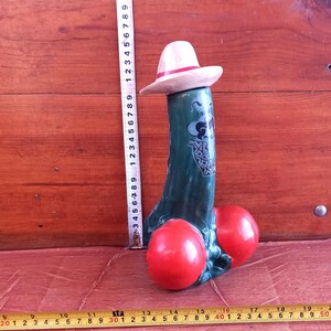 Può includere: Una scultura in ceramica stravagante di una figura stilizzata con corpo verde, globi rossi e cappello bianco con fascia rossa. La figura ha un volto da cartone animato ed è alta circa 23 cm.