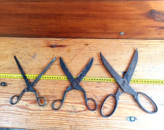 May include: Four vintage scissors with black metal handles and blades, lying on a wooden surface with a yellow measuring tape.