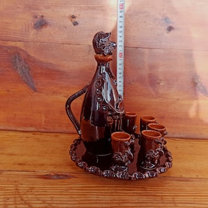 May include: A brown ceramic liquor set with a bottle shaped like a man and six small shot glasses. The set is on a brown wooden surface.