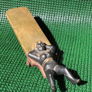 Puede incluir: Un sacabotillas de hierro fundido negro con una figura dorada de una persona reclinada sobre una base de madera. El sacabotillas está sobre una superficie verde.