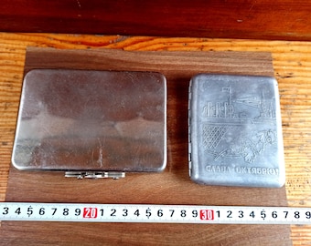 May include: Two silver metal cigarette cases. The case on the left is plain. The case on the right has an inscription that reads "СЛАВА ОКТЯБРЮ!"