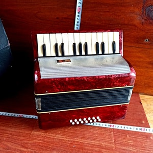 May include: A red and black accordion with a white keyboard and a silver metal plate. The accordion has a decorative pattern on the front and a black bellows.