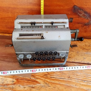 Op de afbeelding: Een vintage zilveren mechanische rekenmachine met een zwarte handgreep en een rij zwarte toetsen. De machine heeft een display met meerdere rijen cijfers. De machine staat op een houten oppervlak.