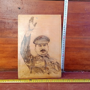 May include: A black and white portrait of a man in a military uniform with a cap. The man is raising his right hand in a salute. The portrait is printed on a wooden plaque.