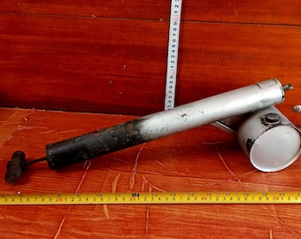 May include: An antique silver-colored air pump with a black handle and a small, round, silver-colored canister. The pump is lying on a wooden surface. A measuring tape is next to the pump.