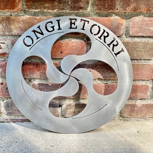 May include: Circular metal wall art with a brushed finish, featuring the text "ONGI ETORRI" and a stylized Basque Lauburu symbol. The artwork is displayed against a weathered brick wall.