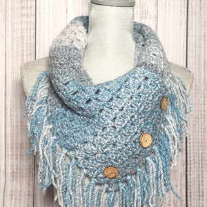 May include: A handmade crochet cowl scarf in shades of blue, gray, and white. The scarf features a fringed edge and is adorned with three wooden buttons. The cowl is displayed on a mannequin, showcasing its texture and design.