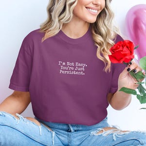 May include: A plum-coloured t-shirt with the text "I'm Not Easy. You're Just Persistent." in white. A red rose is held. The person is wearing ripped blue jeans.