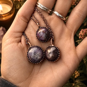 May include: Three round, purple, cabochon-cut stone pendants set in copper bezels with matching copper chains. The stones feature dark, mottled inclusions and a polished, reflective surface. Each pendant is suspended from a delicate copper link chain.