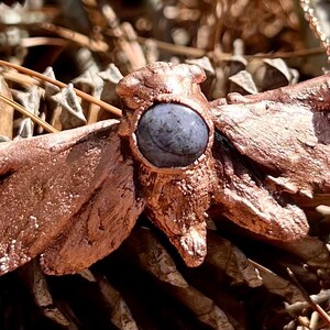 May include: A copper-colored moth pendant necklace with a blue gemstone in the center. The pendant is detailed with textured wings and a body.