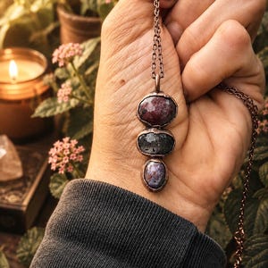 May include: A necklace featuring a copper chain and a pendant with three vertically stacked, raw, irregularly shaped stones in shades of deep red, dark gray, and purple, each encased in a textured copper setting with a rustic finish.