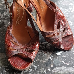 Könnte beinhalten: Ein Paar braune Ledersandalen mit Absatz und Schlangenhautmuster. Die Sandalen haben einen Riemen über dem Fuß und einen Knöchelriemen. Der Markenname "Fly London" ist auf der Innensohle sichtbar.