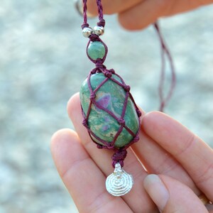 Peut inclure: Un collier composé d'une pierre ovale lisse verte et rose, enserrée dans un filet en cordon macramé violet foncé. Le cordon comporte deux perles argentées au-dessus de la pierre et une petite breloque circulaire argentée texturée en bas.