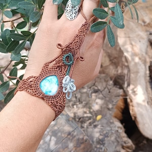 Puede incluir: Una pulsera de mano de macramé marrón con una piedra central de color azul claro en forma de lágrima, intrincados patrones de cordón anudado y un pequeño dije gris en forma de hoja colgando de un lazo de cordón verde oscuro.