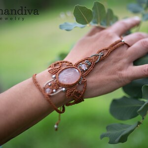 May include: A brown macrame bracelet with a large rose quartz gemstone and smaller clear quartz stones. The bracelet is tied around a hand and finger, creating a finger ring effect. The bracelet is photographed against a green leafy background.
