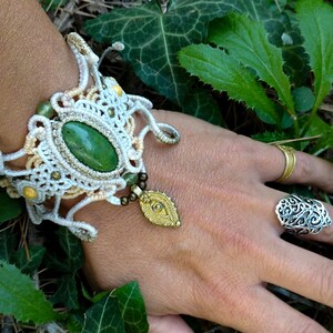 Könnte beinhalten: Ein Makramee-Armband mit einem zentralen ovalen grünen Stein, cremefarbener Kordel sowie kleinen grünen und goldenen Perlen. Ein goldfarbener Metallanhänger hängt am Armband, das zusammen mit einem goldenen Ring und einem silbernen Filigranring getragen wird.
