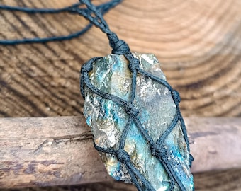 May include: A raw, iridescent stone pendant secured in a black macrame net cage. The stone displays metallic blue, gold, and grey tones with a rough, textured surface. It hangs from a thin, black cord that extends upward from the knotted netting.
