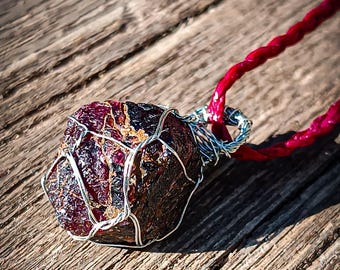 May include: A raw, deep red garnet gemstone pendant encased in a silver-colored wire wrap. The stone features a textured, multifaceted surface with dark inclusions. It is attached to a braided, vibrant red cord necklace that extends to the side.
