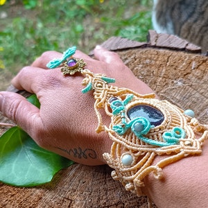 May include: A beige macrame hand ornament featuring intricate knotted patterns, accented with turquoise thread, small white beads, and a central dark purple oval stone. A gold-toned metal component with a small purple gem connects to a finger loop.