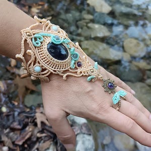 May include: A beige macrame hand ornament featuring a large central dark oval stone, accented with light blue thread details, small round beads, and a metal flower charm with a purple stone that connects to a ring on the middle finger.